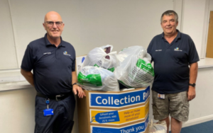 A donation bin filled with bags of donations, two men from The Children's Trust pose alongside it